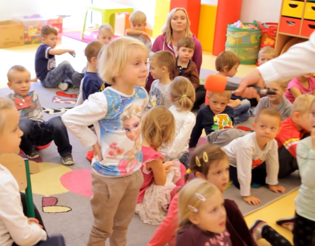 Dzieci kolorowo ubrane siedzą, stoją na dywanie, po prawej stronie widać rękę z mikrofonem, do którego mówi coś chłopczyk